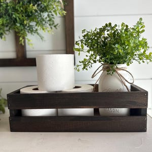 May include: A dark brown wooden crate with three rolls of toilet paper and a white mason jar with green leafy plants. The crate has the word "MASON" printed on the front.