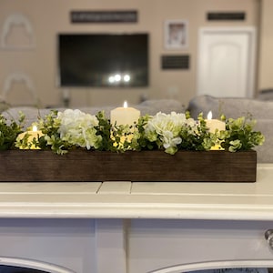 Puede incluir: Un centro de mesa de caja de madera con flores de hortensia blancas y vegetaci&oacute;n, decorado con velas encendidas.