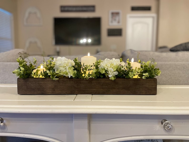 Puede incluir: Un centro de mesa de caja de madera con flores de hortensia blancas y vegetaci&oacute;n, decorado con velas encendidas.