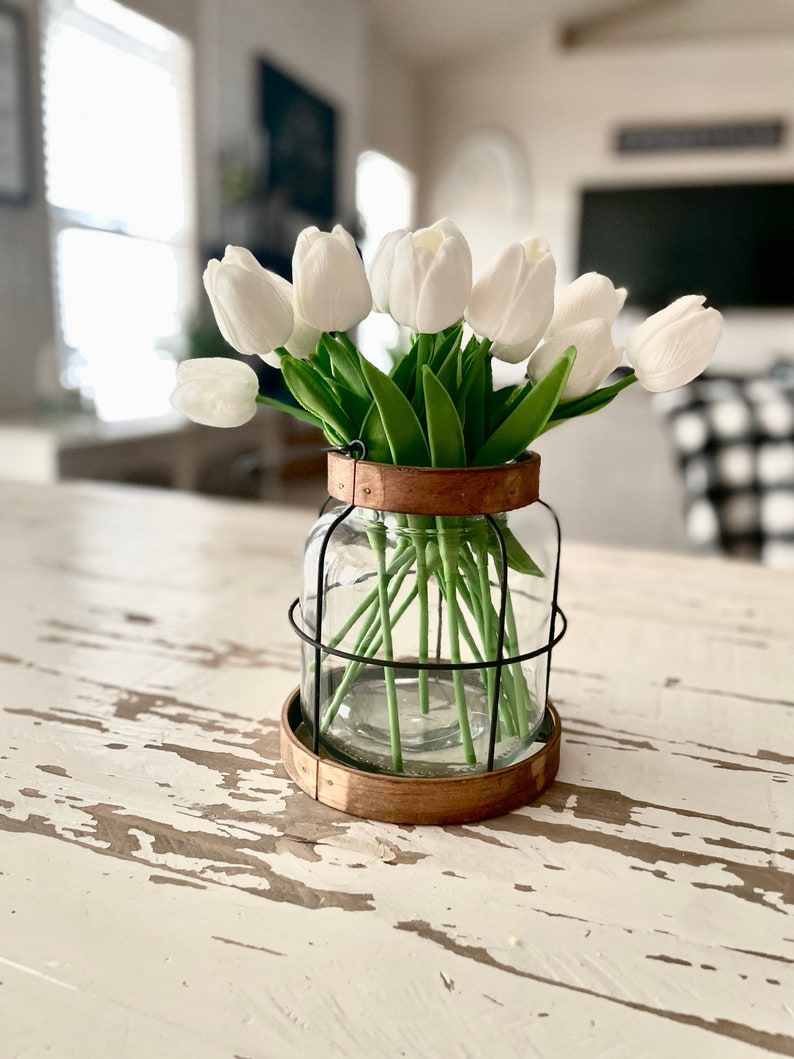 K&ouml;nnte beinhalten: Wei&szlig;e Tulpen in einer klaren Glasvase mit einem schwarzen Drahtk&auml;fig und einem Holzsockel. Die Vase steht auf einem wei&szlig; gestrichenen Holztisch.