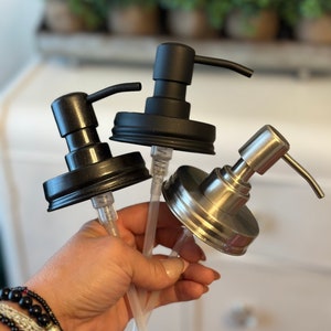May include: Three soap dispensers with black and silver pump tops and clear tubes. The dispensers are made from glass jars with black and silver lids.