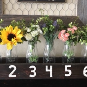 Op de afbeelding: Een houten doos met zes glazen melkflessen met verschillende soorten bloemen. De doos is zwart geverfd met witte nummers 1 tot en met 6.