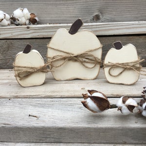 May include: Three white wooden pumpkins with brown stems and twine tied around them. The pumpkins are arranged in a row on a wooden surface.