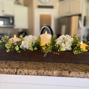 Puede incluir: Un centro de mesa de caja de madera con tres velas blancas de pilar, flores de hortensia blancas y ramitas verdes.