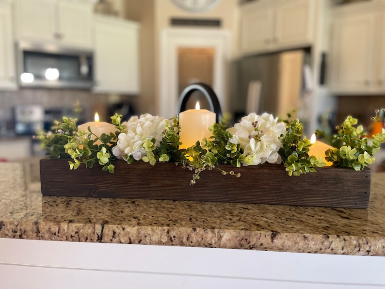 Puede incluir: Un centro de mesa de caja de madera con tres velas blancas de pilar, flores de hortensia blancas y ramitas verdes.
