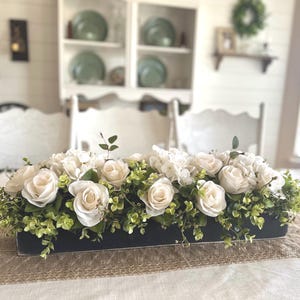 May include: A white flower arrangement in a black rectangular planter. The arrangement features white roses and hydrangeas with green foliage.