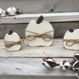 May include: Three white wooden pumpkins tied with twine, arranged on a wooden surface with cotton bolls.
