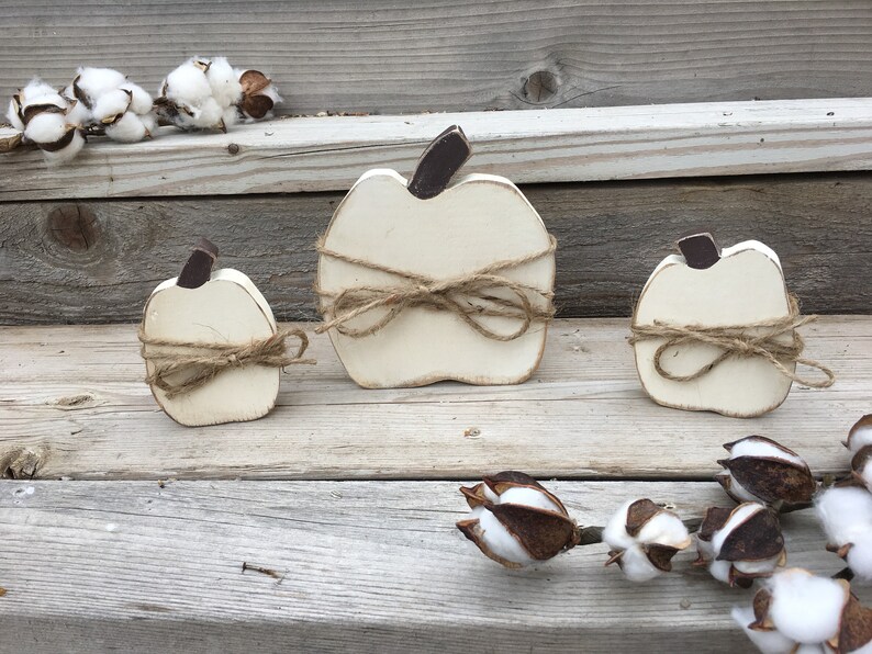 May include: Three white wooden pumpkins tied with twine, arranged on a wooden surface with cotton bolls.