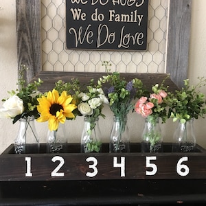 K&ouml;nnte beinhalten: Eine Holzkiste mit sechs Glasmilchflaschen, jede mit einer anderen Blumenarrangement. Die Blumen umfassen wei&szlig;e Rosen, Sonnenblumen, wei&szlig;e Blumen, Lavendel, rosa Rosen und Gr&uuml;n. Die Kiste ist schwarz lackiert mit wei&szlig;en Zahlen von 1 bis 6.