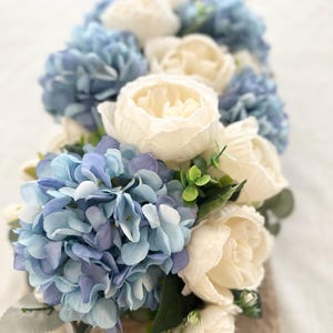 K&ouml;nnte beinhalten: Nahaufnahme eines Blumenarrangements mit blauen Hortensien und cremefarbenen Pfingstrosen. Das Arrangement umfasst gr&uuml;nes Laub und ist auf einer neutralen Oberfl&auml;che platziert. Die Farbpalette erzeugt eine weiche und elegante &Auml;sthetik.