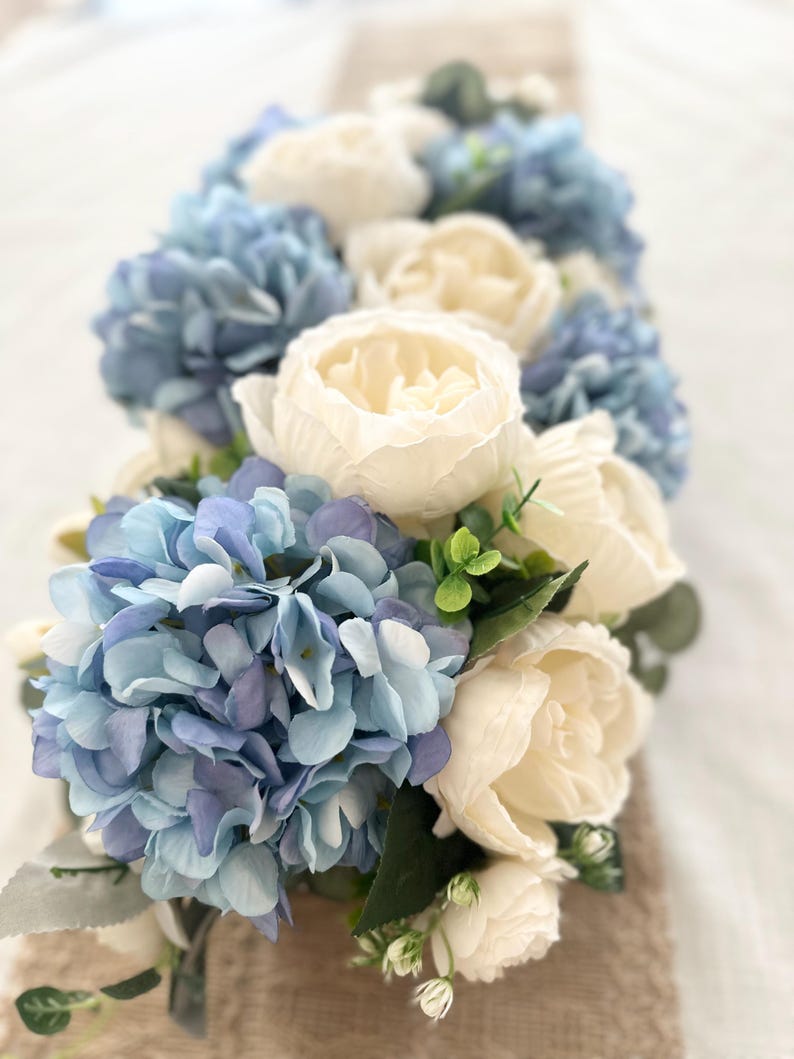 K&ouml;nnte beinhalten: Nahaufnahme eines Blumenarrangements mit blauen Hortensien und cremefarbenen Pfingstrosen. Das Arrangement umfasst gr&uuml;nes Laub und ist auf einer neutralen Oberfl&auml;che platziert. Die Farbpalette erzeugt eine weiche und elegante &Auml;sthetik.