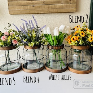 K&ouml;nnte beinhalten: Vier Glasvasen mit Drahtk&auml;figen, gef&uuml;llt mit verschiedenen Blumenarrangements. Die Vasen haben Holzsockel und sind mit "BLEND 5", "BLEND 4", "WHITE TULIPS" und "BLEND 2" beschriftet.
