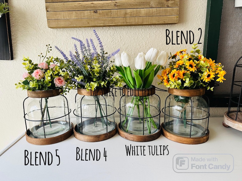 K&ouml;nnte beinhalten: Vier Glasvasen mit Drahtk&auml;figen, gef&uuml;llt mit verschiedenen Blumenarrangements. Die Vasen haben Holzsockel und sind mit "BLEND 5", "BLEND 4", "WHITE TULIPS" und "BLEND 2" beschriftet.