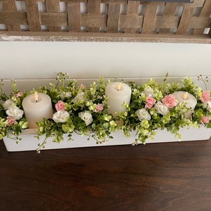 May include: A white rectangular centerpiece with three lit pillar candles surrounded by greenery, white, and pink flowers. The arrangement sits on a dark wooden surface, creating a contrast with the bright floral display. A decorative wooden element is in the background.