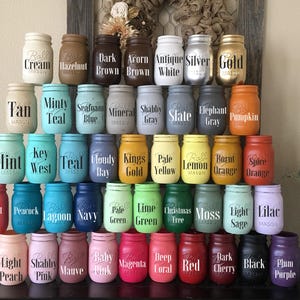 May include: A collection of 48 mason jars painted in various colors with different text labels. The jars are arranged in rows, with each row featuring a different color theme. The jars are all the same size and shape, and they are all made of glass.