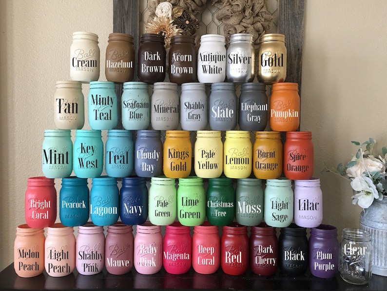 May include: A collection of 48 mason jars painted in various colors with different text labels. The jars are arranged in rows, with each row featuring a different color theme. The jars are all the same size and shape, and they are all made of glass.