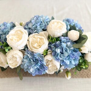K&ouml;nnte beinhalten: Ein Blumenarrangement mit wei&szlig;en Pfingstrosen und blauen Hortensien. Die Blumen sind mit gr&uuml;nem Laub arrangiert und vor einem neutralen Hintergrund platziert. Das Arrangement ist eine Mischung aus Texturen und Farben.