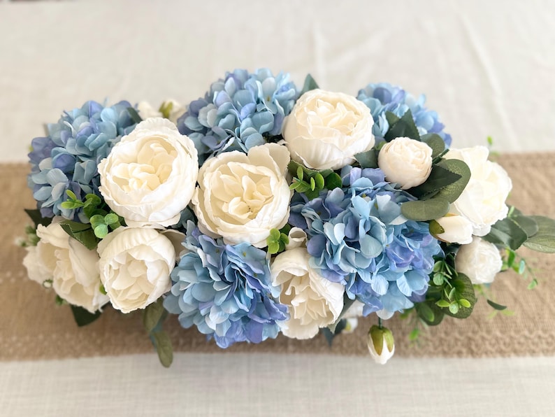 K&ouml;nnte beinhalten: Ein Blumenarrangement mit wei&szlig;en Pfingstrosen und blauen Hortensien. Die Blumen sind mit gr&uuml;nem Laub arrangiert und vor einem neutralen Hintergrund platziert. Das Arrangement ist eine Mischung aus Texturen und Farben.