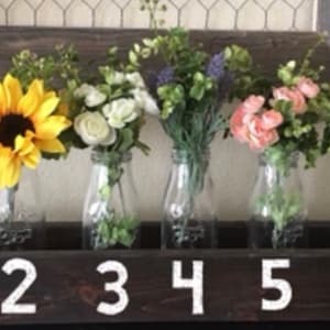 May include: A wooden box with six glass milk bottles, each holding a different type of flower arrangement. The bottles are numbered 1 through 6 on the front of the box.