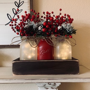 May include: Three mason jars with frosted glass and fairy lights inside, filled with red berry sprigs and pine cones, sitting in a dark brown wooden box.