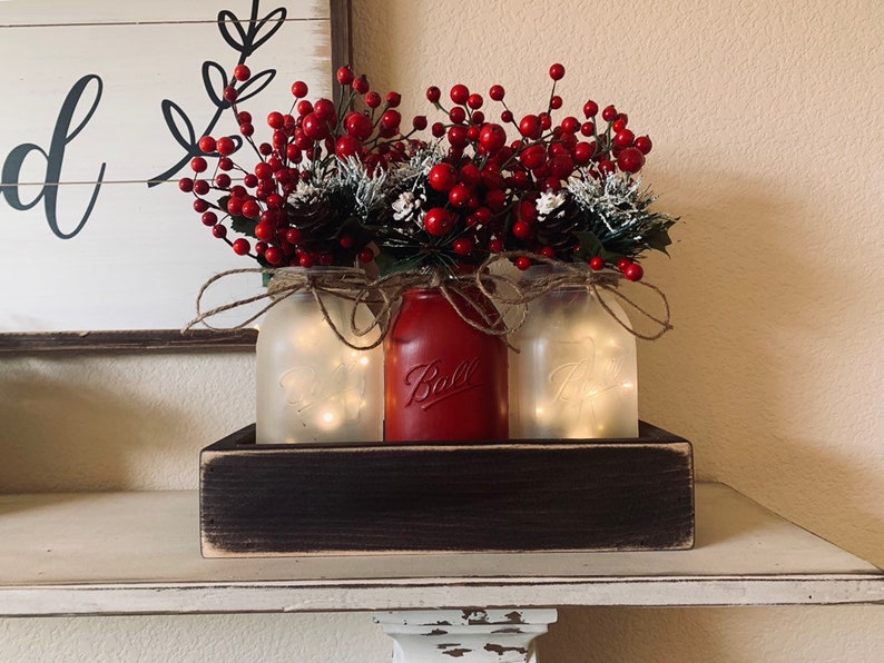 May include: Three mason jars with frosted glass and fairy lights inside, filled with red berry sprigs and pine cones, sitting in a dark brown wooden box.