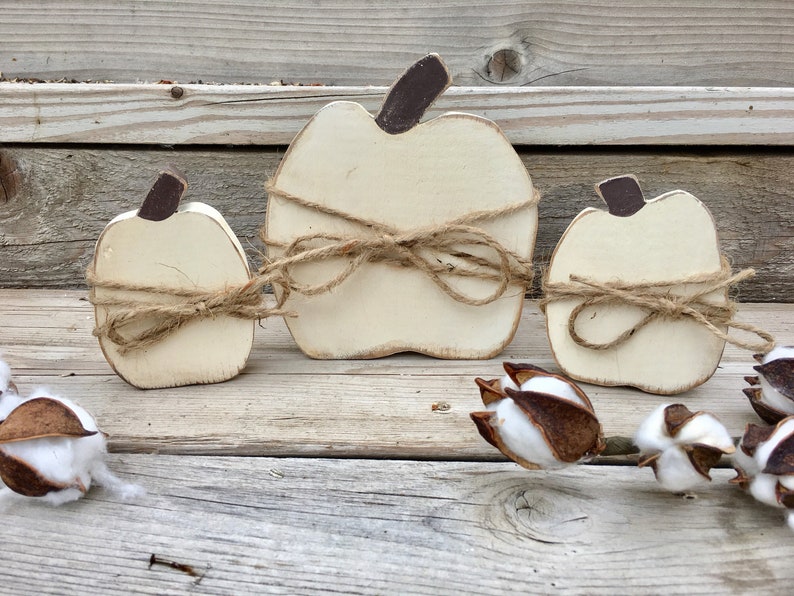 May include: Three white wooden pumpkins with brown stems, tied with twine, arranged on a wooden surface.