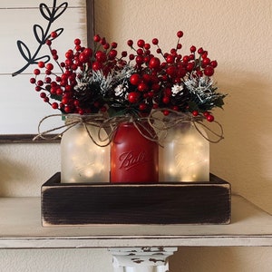 May include: Three mason jars with red berries and pine cones, lit with white fairy lights, in a rustic wooden box.