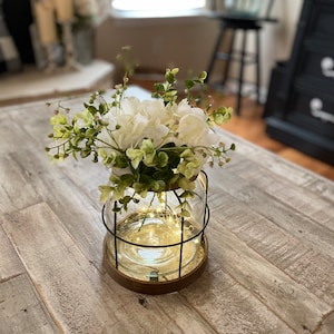 May include: A white floral arrangement in a clear glass jar with a wire cage and string lights. The jar is sitting on a wooden table.