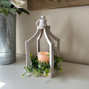 May include: White wooden lantern with a white candle inside and green foliage around the base.