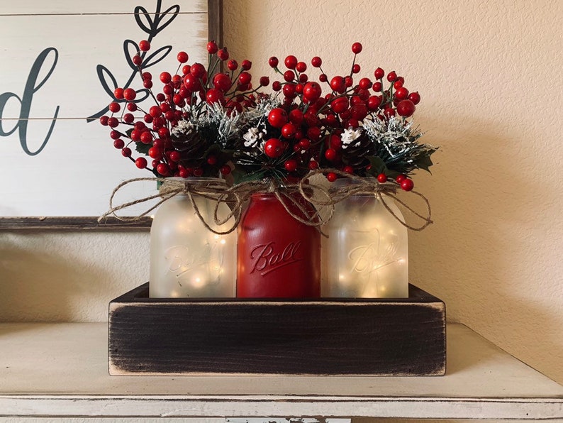 May include: Three mason jars with red berry sprigs and pine cones, lit with fairy lights, in a dark wood box.