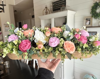May include: A wooden bowl filled with a floral arrangement of pink, white, and peach flowers. The flowers are surrounded by green leaves.
