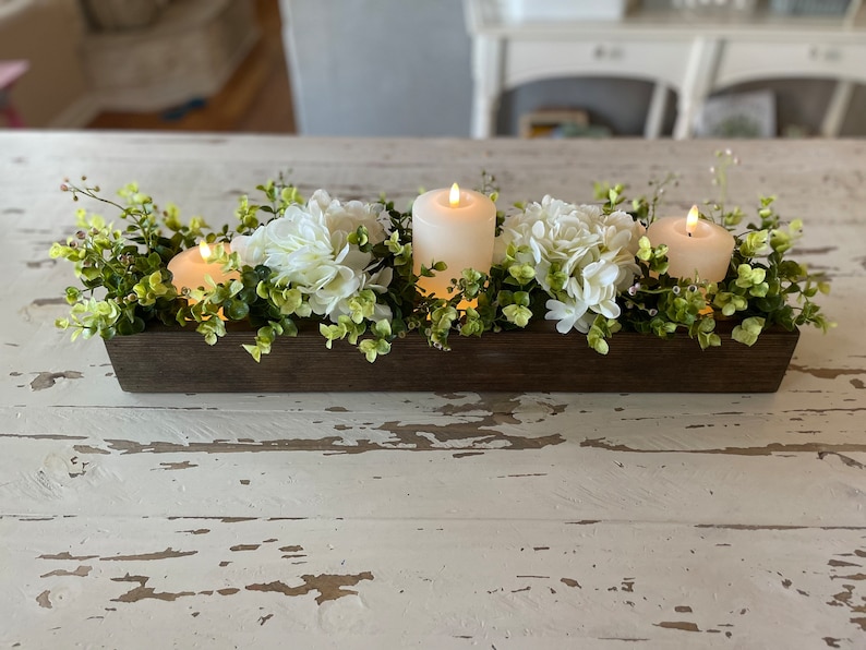 Puede incluir: Una caja de madera como centro de mesa con flores blancas de hortensia y tres velas blancas cil&iacute;ndricas. La caja est&aacute; llena de follaje verde.