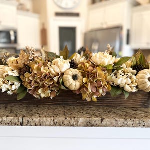 May include: A wooden box centerpiece filled with fall-themed floral arrangements. The arrangement includes cream-colored peonies, orange hydrangeas, small pumpkins, and pinecones. The flowers are arranged in a rectangular, dark brown wooden box.