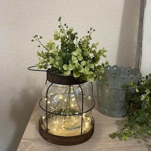 K&ouml;nnte beinhalten: Ein klares Glasgef&auml;&szlig; mit einem Metallk&auml;fig drumherum, gef&uuml;llt mit gr&uuml;nen, bl&auml;ttrigen Pflanzen und einer Lichterkette mit wei&szlig;en Lichtern. Das Gef&auml;&szlig; steht auf einer Holzoberfl&auml;che.