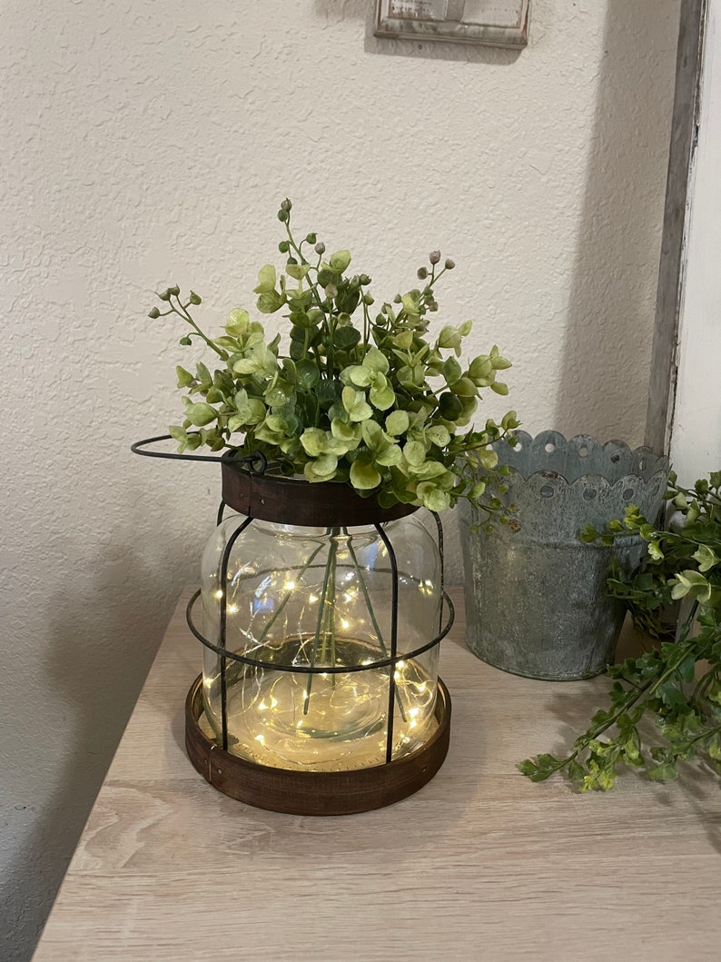 K&ouml;nnte beinhalten: Ein klares Glasgef&auml;&szlig; mit einem Metallk&auml;fig drumherum, gef&uuml;llt mit gr&uuml;nen, bl&auml;ttrigen Pflanzen und einer Lichterkette mit wei&szlig;en Lichtern. Das Gef&auml;&szlig; steht auf einer Holzoberfl&auml;che.