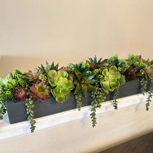 May include: A long, rectangular gray planter filled with a variety of green and brown succulents. The planter has a rustic look and the succulents are spilling over the edges.
