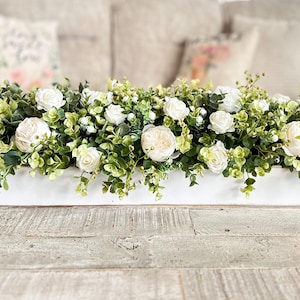 May include: A white wooden tray filled with a floral arrangement of white roses and green foliage.