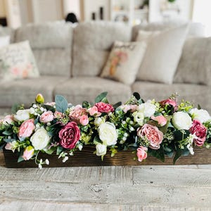May include: A wooden box filled with a floral arrangement of pink, white, and blush-colored flowers. The flowers are arranged in a long, rectangular box and are surrounded by greenery.