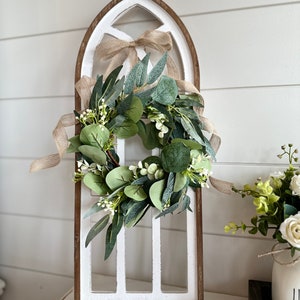 May include: A white wooden arched window frame with a green eucalyptus wreath with white flowers and a brown burlap ribbon. The wreath is hanging on the window frame.