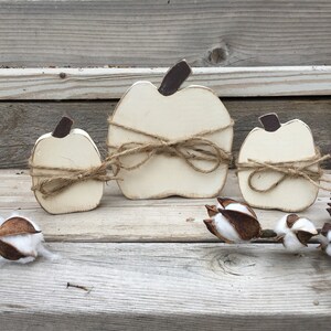 May include: Three white wooden pumpkins of varying sizes, each tied with twine and a bow. The pumpkins are arranged on a wooden surface with cotton bolls.