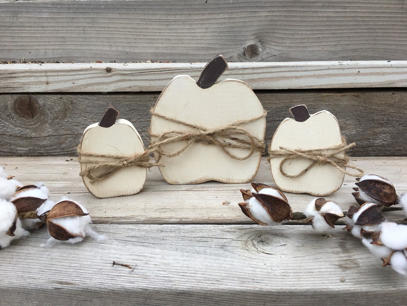 May include: Three white wooden pumpkins of varying sizes, each tied with twine and a bow. The pumpkins are arranged on a wooden surface with cotton bolls.