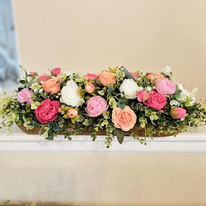 May include: A floral arrangement in a wooden trough. The arrangement features a variety of roses in shades of pink, peach, and white, interspersed with green foliage. The trough is rectangular and sits on a white surface.