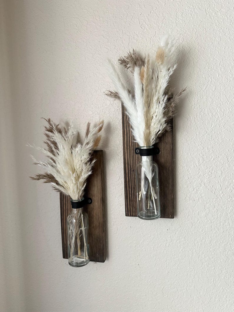 May include: Two wooden wall mounted vases with clear glass bottles and dried pampas grass in shades of white and beige.