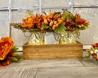 May include: A wooden box with three mason jars filled with fall-colored flowers and lights. The jars are wrapped with twine and the box has a rustic finish. The flowers are orange, red, and yellow.
