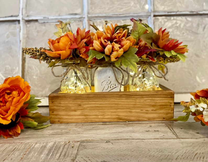 Fall centerpiece featuring two clear jars with lights giving off a soft glow and one white jar. Each jar holds vibrant fall floral arrangements with autumn leaves and sunflowers. The jars are set in a handmade lightly stained rustic box.