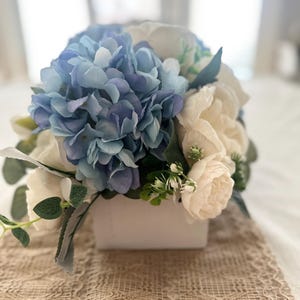 K&ouml;nnte beinhalten: Ein Blumenarrangement mit blauen Hortensien und cremefarbenen Rosen in einer wei&szlig;en quadratischen Vase. Gr&uuml;ne Bl&auml;tter und Akzente erg&auml;nzen den nat&uuml;rlichen Look. Das Arrangement steht auf einem beigen Spitzentischl&auml;ufer.