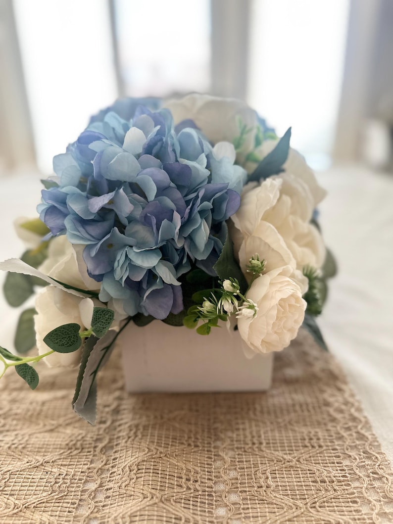 K&ouml;nnte beinhalten: Ein Blumenarrangement mit blauen Hortensien und cremefarbenen Rosen in einer wei&szlig;en quadratischen Vase. Gr&uuml;ne Bl&auml;tter und Akzente erg&auml;nzen den nat&uuml;rlichen Look. Das Arrangement steht auf einem beigen Spitzentischl&auml;ufer.