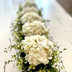 May include: A white farmhouse style centerpiece with a row of white hydrangea blooms and greenery in a black planter.