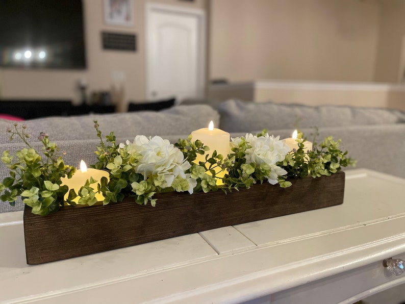Puede incluir: Una caja de madera como centro de mesa con tres velas blancas y flores blancas. La caja es marr&oacute;n y tiene un acabado r&uacute;stico. Las flores son blancas y verdes y est&aacute;n dispuestas alrededor de las velas.