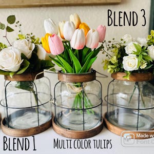 K&ouml;nnte beinhalten: Drei Glasvasen mit Drahtk&auml;figen, jede gef&uuml;llt mit verschiedenen Blumenarrangements. Die erste Vase enth&auml;lt wei&szlig;e Rosen und Gr&uuml;nzeug, die zweite enth&auml;lt mehrfarbige Tulpen und die dritte enth&auml;lt wei&szlig;e Rosen und Gr&uuml;nzeug. Die Vasen sind mit "BLEND 1", "MULTI COLOR TULIPS" und "BLEND 3" beschriftet.